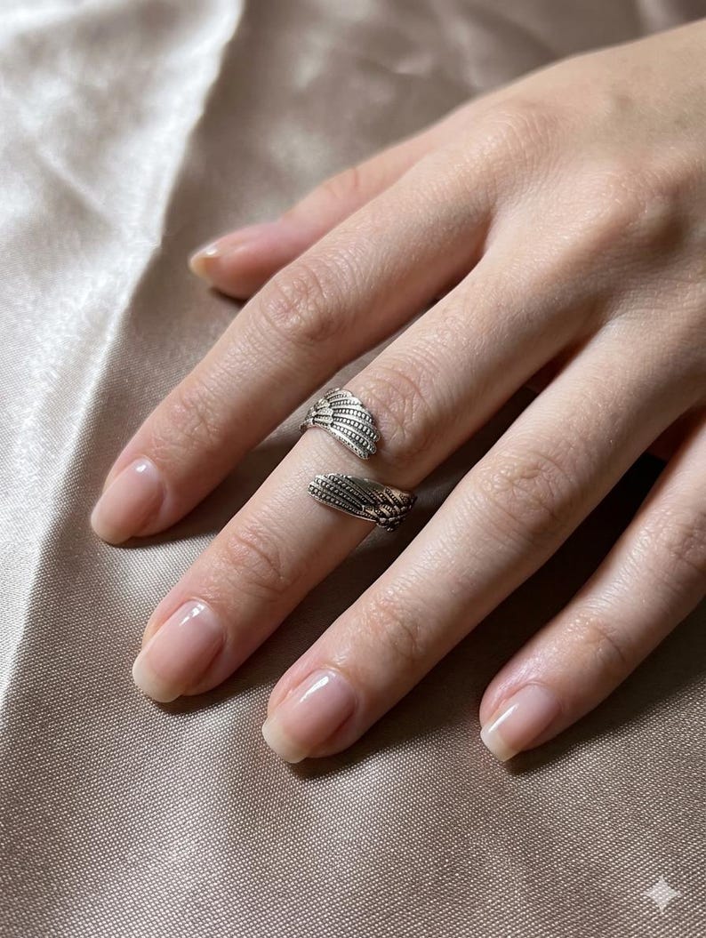 May include: Two silver rings, each featuring a detailed wing design, are worn on a hand. The rings are on the index and middle fingers. The nails are neatly manicured with a natural, glossy finish. The background is a soft, neutral color.