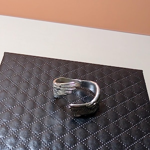May include: A silver-colored cuff bracelet with a wing design. The bracelet is open and has detailed feather-like patterns. It rests on a black, diamond-patterned surface.