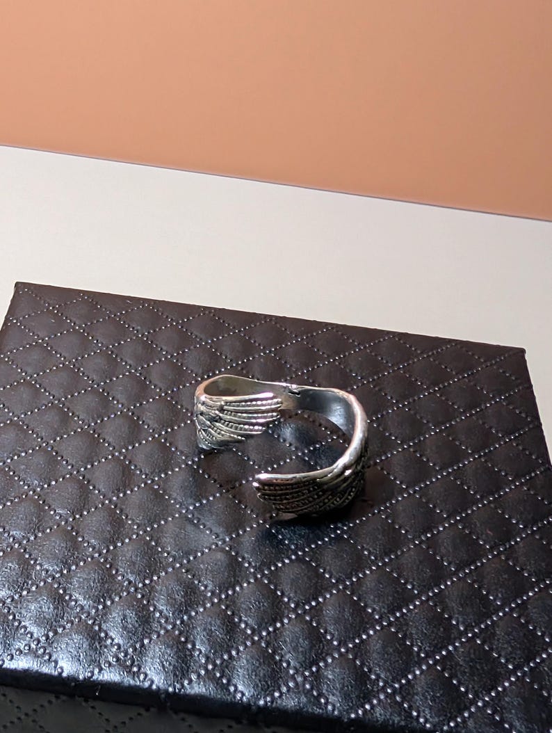 May include: A silver-colored cuff bracelet with a wing design. The bracelet is open and has detailed feather-like patterns. It rests on a black, diamond-patterned surface.