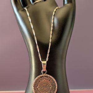 May include: A silver-toned circular pendant featuring an intricate embossed Aztec calendar design, suspended from a delicate twisted silver-toned chain. The pendant has a polished metallic finish with dark recessed details highlighting the pattern.
