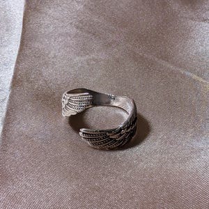 May include: A silver-colored ring with a wing design. The ring features detailed feather patterns and is set against a neutral background. The ring is open-ended, allowing for size adjustment.