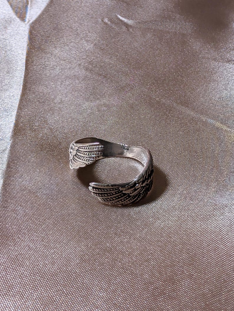 May include: A silver-colored ring with a wing design. The ring features detailed feather patterns and is set against a neutral background. The ring is open-ended, allowing for size adjustment.