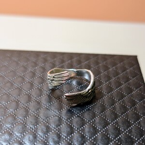 May include: A silver-colored ring with an open design. The ring features wing-like details on each end, with intricate feather patterns. The ring is displayed on a black, textured surface.