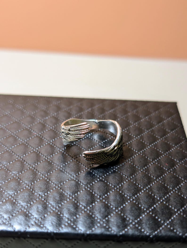 May include: A silver-colored ring with an open design. The ring features wing-like details on each end, with intricate feather patterns. The ring is displayed on a black, textured surface.