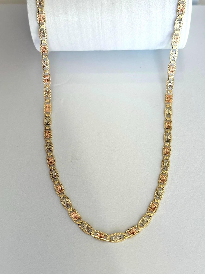 Op de afbeelding: Een goudkleurige ketting met een herhalend patroon van stervormige elementen en ovale schakels. De ketting heeft een driekleurig ontwerp, met goud-, ros&eacute;goud- en zilvertinten. De ketting wordt gepresenteerd tegen een witte achtergrond.