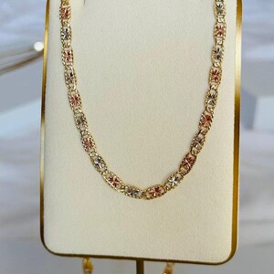 Op de afbeelding: Een gouden ketting met een herhalend bloemmotief. De ketting heeft afwisselende secties van goud, ros&eacute;goud en zilverkleurig metaal. De ketting wordt gepresenteerd op een witte standaard met een gouden rand.