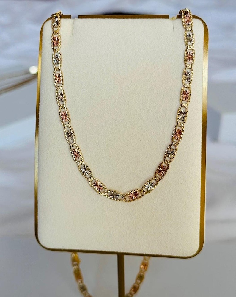 Op de afbeelding: Een gouden ketting met een herhalend bloemmotief. De ketting heeft afwisselende secties van goud, ros&eacute;goud en zilverkleurig metaal. De ketting wordt gepresenteerd op een witte standaard met een gouden rand.
