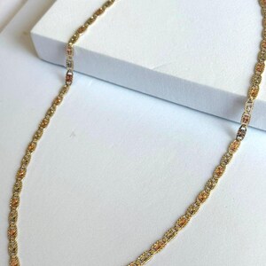 Op de afbeelding: Een goudkleurige ketting met een delicaat, ingewikkeld ontwerp. De ketting heeft afwisselende secties van goud, ros&eacute;goud en een lichtere goudtint, waardoor een meerkleurig effect ontstaat. De ketting wordt op een wit oppervlak getoond.
