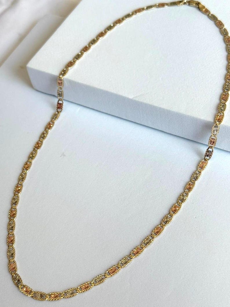 Op de afbeelding: Een goudkleurige ketting met een delicaat, ingewikkeld ontwerp. De ketting heeft afwisselende secties van goud, ros&eacute;goud en een lichtere goudtint, waardoor een meerkleurig effect ontstaat. De ketting wordt op een wit oppervlak getoond.