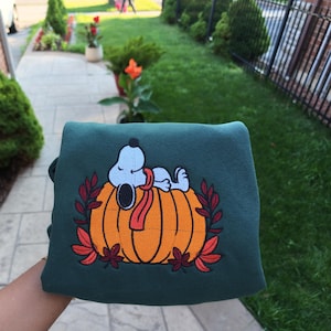 May include: A dark green sweatshirt featuring an embroidered graphic of the cartoon character Snoopy wearing a red scarf and resting on top of a large orange pumpkin, surrounded by a decorative border of red and dark orange autumn leaves.
