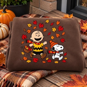 Peut inclure: Un sweat-shirt marron avec une impression graphique de Charlie Brown et Snoopy courant parmi des feuilles d'automne. Charlie Brown porte un t-shirt jaune à zigzag noir, une écharpe rouge et orange, et tient un panier. Snoopy porte un bonnet orange.