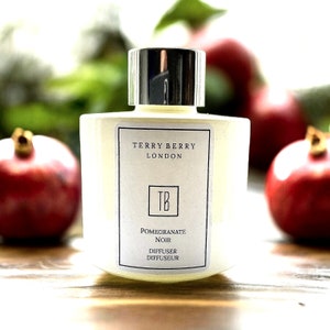 Puede incluir: Una botella blanca con una tapa plateada y una etiqueta que dice "Terry Berry London" y "Pomegranate Noir Diffuser Diffuseur". La botella está sobre una superficie de madera con dos granadas rojas en el fondo.