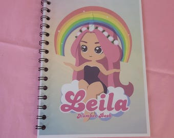 Può includere: Un quaderno a spirale con un arcobaleno colorato e una ragazza dei cartoni animati con i capelli rosa che indossa una corona di fiori. La copertina del quaderno dice "Leila Number Book".