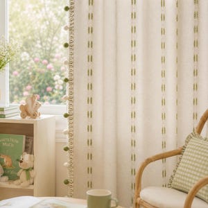 May include: Cream-colored curtains with vertical green stitch detailing and a pom-pom trim. A wicker chair with a green and white checkered pillow sits beside the curtains. A green mug and open book are on a table. A shelf holds books and stuffed animals.