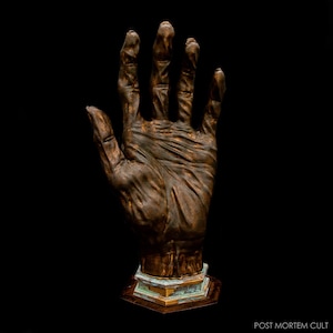 Puede incluir: Escultura de bronce de una mano humana, con la palma hacia arriba, sobre un fondo negro. La mano, con detalles de la piel, está montada sobre una base geométrica. El texto "POST MORTEM CULT" es visible.