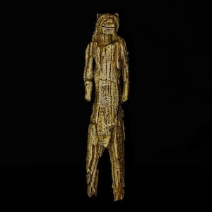 Könnte beinhalten: Eine geschnitzte Elfenbeinfigur einer menschlichen Gestalt mit einem bärenartigen Kopf. Die Figur steht aufrecht mit den Armen an den Seiten. Die Figur ist mit aufwendigen Schnitzereien verziert und hat ein verwittertes, altes Aussehen.