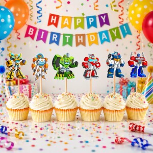 May include: Six cupcakes topped with robot character paper cutouts on wooden picks. Each cupcake features white frosting in a paper liner. The background displays a colorful Happy Birthday banner, balloons, and confetti on a white surface.