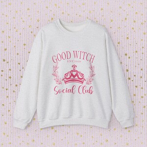 Puede incluir: Una sudadera gris jaspeada con un diseño gráfico rosa que dice "Good Witch Est. 1939 Social Club" con una corona y detalles florales.