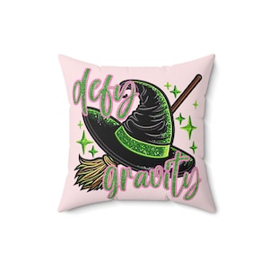 Peut inclure: Un coussin rose avec un chapeau de sorcière vert et un balai, et le texte "defy gravity" en paillettes roses.