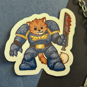 Puede incluir: Un gato de dibujos animados con armadura azul y dorada y sosteniendo una espada roja y plateada. El gato tiene pelaje naranja y una pajarita amarilla.