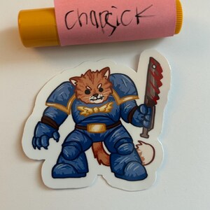 Puede incluir: Un gato de dibujos animados azul y naranja con armadura y sosteniendo una espada ensangrentada. El gato tiene una expresi&oacute;n feroz en su rostro. La pegatina est&aacute; sobre un fondo blanco.
