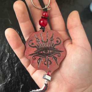 Pode incluir: Um porta-chaves de madeira com um pendente circular castanho-avermelhado com bordas recortadas e um design de olho gravado a preto. Inclui um anel de metal prateado, uma conta de vidro craquelado vermelho, uma conta cúbica prateada e uma borla multicolorida.