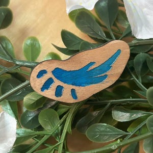 May include: A small, irregularly shaped wooden pin featuring a smooth, light brown natural wood grain finish. The surface is decorated with a painted design in a vibrant, glossy blue, consisting of a central wing-like shape and four small dots.