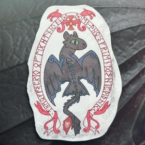 May include: A sticker depicting a black dragon with blue wing details and green eyes, encircled by a red and white rune-inscribed border. The dragon has a long tail with a red tag. The sticker is on a dark grey surface.
