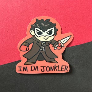 Puede incluir: Una pegatina de dibujos animados con un personaje con abrigo marrón, máscara blanca y sosteniendo un cuchillo. La pegatina tiene el texto "IM DA JONKLER" en letras negras sobre un fondo rojo. La pegatina está sobre una superficie roja y negra.