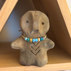 May include: A small, tan-colored clay figurine with a stylized human form. The figurine has a simple face with two eye holes and a necklace of blue, yellow, and tan beads. The figure is displayed on a wooden shelf.