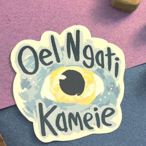 Puede incluir: Una pegatina con el texto "Oel Ngati Kameie" en azul oscuro, que presenta un ojo estilizado con acentos amarillos y azules. La pegatina está sobre una superficie azul y morada.