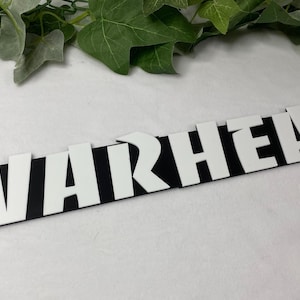May include: A black and white sign that reads "WARHEAD" in bold, sans-serif font. The letters are white and stand out against the black background. The sign is rectangular with rounded edges.