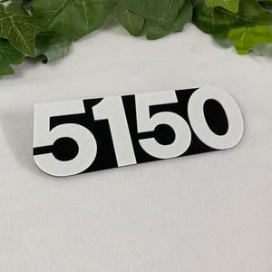 May include: A rectangular address plaque with the number "5150" in white, set against a black background. The numbers are bold and stylized. The plaque is displayed on a white surface, with green foliage in the background.