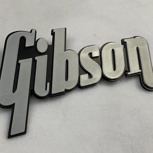 Op de afbeelding: Een driedimensionaal bord met het woord Gibson in een gestileerd scriptlettertype. De belettering bestaat uit een matte zilveren toplaag met een textuur, gemonteerd op een solide zwarte basislaag die de contouren van de letters volgt.