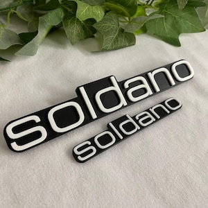 Puede incluir: Dos emblemas rectangulares negros con la palabra "Soldano" en blanco, uno grande y otro pequeño. Los emblemas están sobre una superficie blanca, con hojas verdes en el fondo.