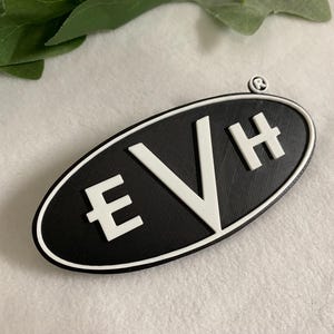 Könnte beinhalten: Schwarz-weißes ovales Emblem mit den weißen Buchstaben "EVH". Das Emblem hat einen schwarzen, strukturierten Hintergrund und einen weißen Rand. Oben befindet sich ein kleines, kreisförmiges Symbol.
