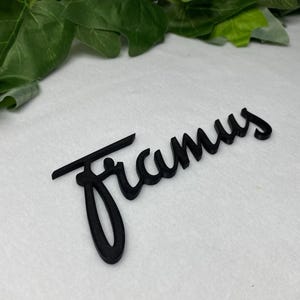 May include: Black 3D-printed name sign with the name "Framus" in a cursive font. The sign is placed on a white surface, with green leaves in the background. The sign is a decorative item.