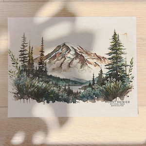 Close up of an unframed Mount Rainier National Park watercolor painting poster on a wooden desk, illuminated by natural sunlight and dappled leaf shadows.