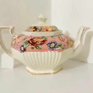 May include: A ceramic teapot featuring a pink upper section decorated with a colorful floral pattern of red, purple, and yellow flowers with green leaves. The lower portion is cream-colored with vertical fluting and gold trim on the edges.
