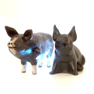 May include: Two pig figurines. The left pig is standing, coloured black with white patches on its face and legs. The right pig is sitting, coloured solid black. Both figurines feature pointed ears and snout details, presented against a plain white background.