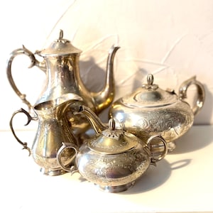 May include: A silver-plated tea service set consisting of a tall teapot, a lidded sugar bowl, a creamer, and a lidded waste bowl. Each piece features intricate floral engravings, curved handles, and ornate finials on the lids of the vessels.