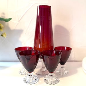 May include: A tall, tapered ruby red glass vase positioned behind four matching ruby red glass goblets. Each goblet features a clear, textured glass base with a distinct hobnail or bubble pattern, providing a contrast to the smooth red bowls.