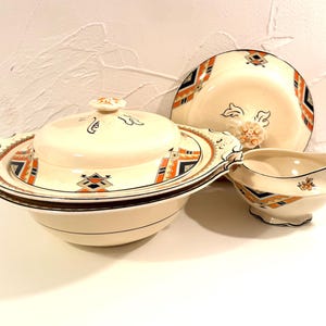 Può includere: Un set da portata in ceramica color crema composto da una zuppiera con coperchio e una salsiera. Entrambi i pezzi sono decorati con motivi geometrici in arancione, nero e blu, arricchiti da piccoli motivi floreali e bordi marrone scuro.