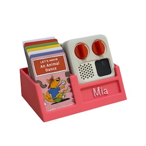 May include: A pink card holder with a white and orange device. The holder contains a stack of colourful cards with the text "LET'S MOVE An Animal Dance". The device has two orange knobs and the name "Mia" on the holder.