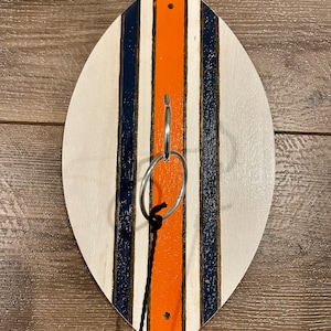 Puede incluir: Un juego de lanzamiento de anillos de madera en forma de tabla de surf blanca con rayas azules y naranjas. Un anillo de metal está colgado de una cuerda negra.