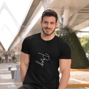 Op de afbeelding: Een man draagt een zwart T-shirt met het woord "Pray" in witte cursieve letters. Hij staat met een gebogen been en zijn voet rust op een betonnen pilaar. Hij draagt een zwarte broek en witte sneakers.