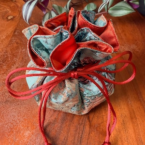 May include: A small, drawstring pouch with a teal and gold brocade exterior and a bright red interior. The pouch is gathered at the top and tied with a red cord. The pouch is likely used for jewelry or small gifts.