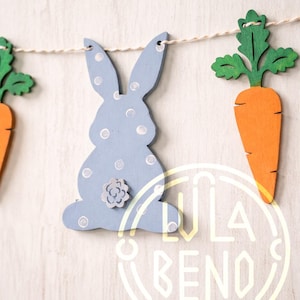 May include: A wooden Easter garland featuring a light blue bunny with white polka dots and a flower detail, along with orange carrots with green tops. The garland is strung on a natural twine, perfect for Easter decorations.