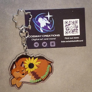 May include: A clear acrylic keychain with a cartoon fox and sunflower design. The keychain has a silver-coloured flower-shaped clasp. The text "Hidden Disability" is on the keychain. A business card is behind the keychain.