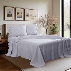 May include: A bed is made with a light purple sheet set. The set includes a flat sheet, fitted sheet, and two pillowcases. The pillowcases and the top sheet have a ruffled edge. The bed is in a bedroom with artwork and a large window.